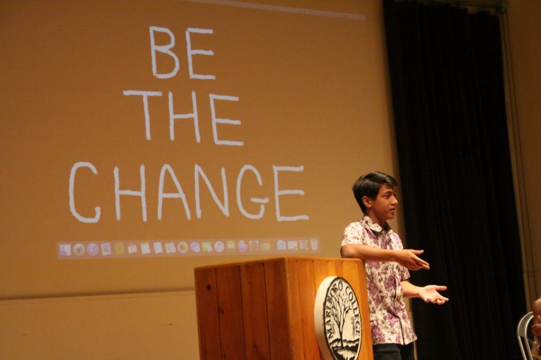 Student talking on podium with projection behind that reads, "Be the Change"