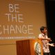 Student talking on podium with projection behind that reads, "Be the Change"